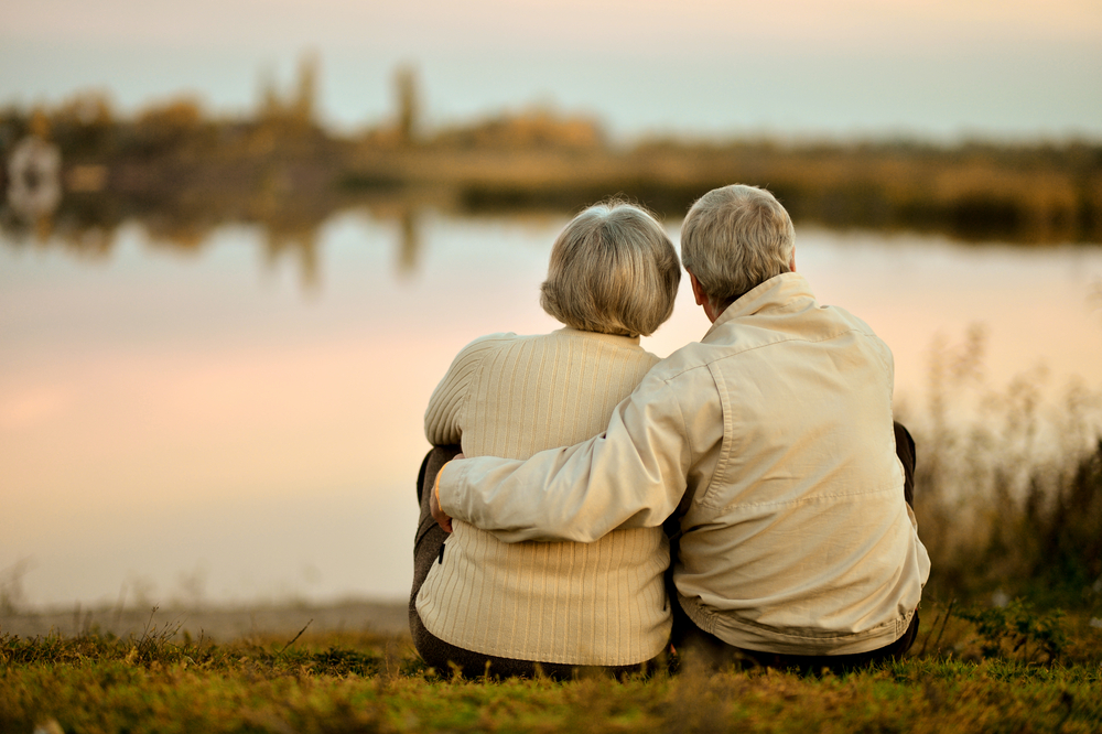 Retired elderly couple sat by lake thinking about their pensions
