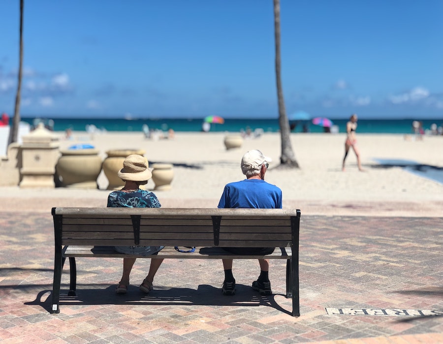 An elderly couple sitting on a bench looking at a sunny beach